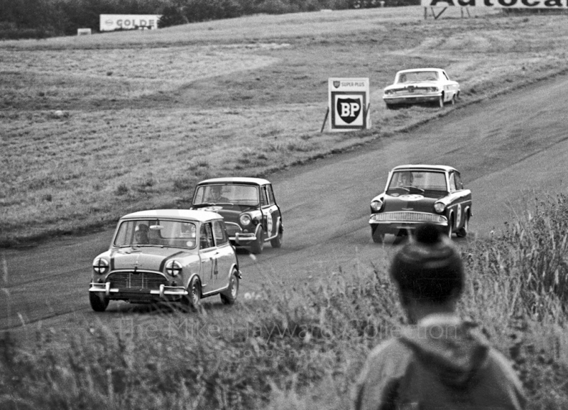 Edward Savory, Don Moore Mini Cooper S; Harry Ratcliffe, Vitafoam Mini Cooper S; and Mike Young, Superspeed Ford Anglia; Oulton Park, Gold Cup meeting 1964. Parked on the grass is the Ford Galaxie of Jack Sears, who retired with ignition failure.
